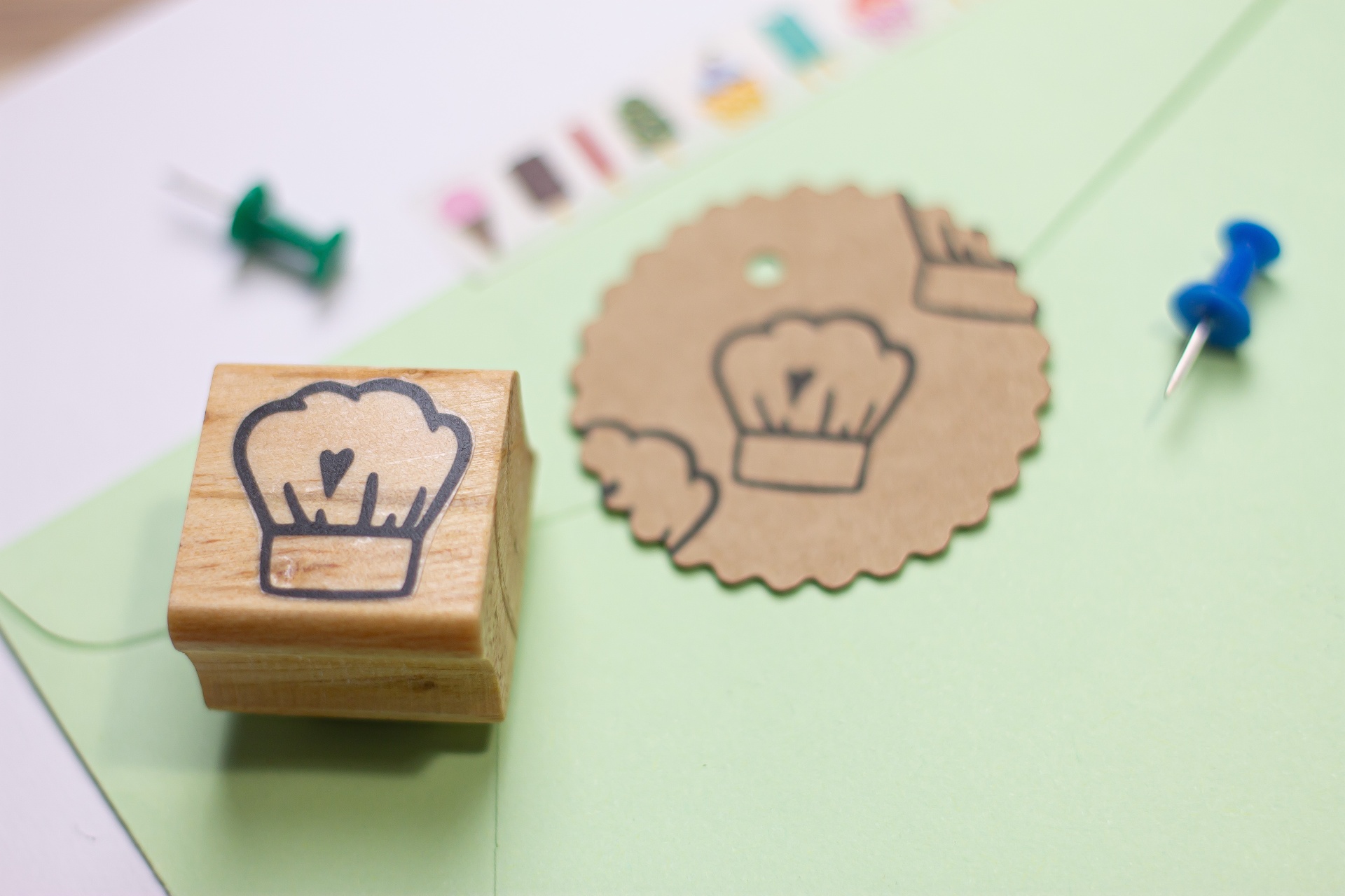 Selo de carimbo de madeira com desenho de chapéu de chef e papel em forma de selo ao lado de envelope verde e alfinetes