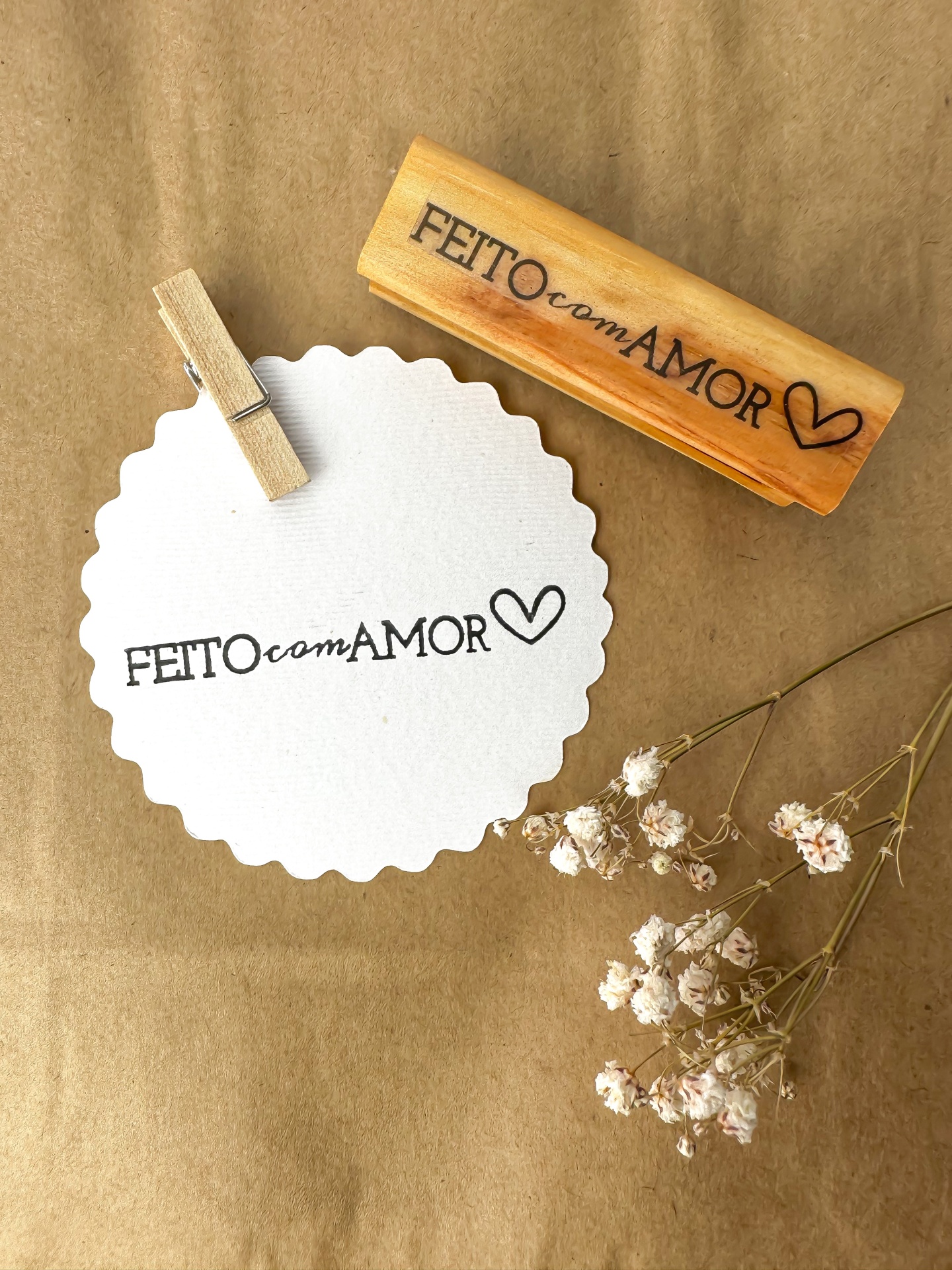 Carimbo de madeira com texto 'FEITO com AMOR' e etiqueta branca com o mesmo texto, com flores secas ao lado.