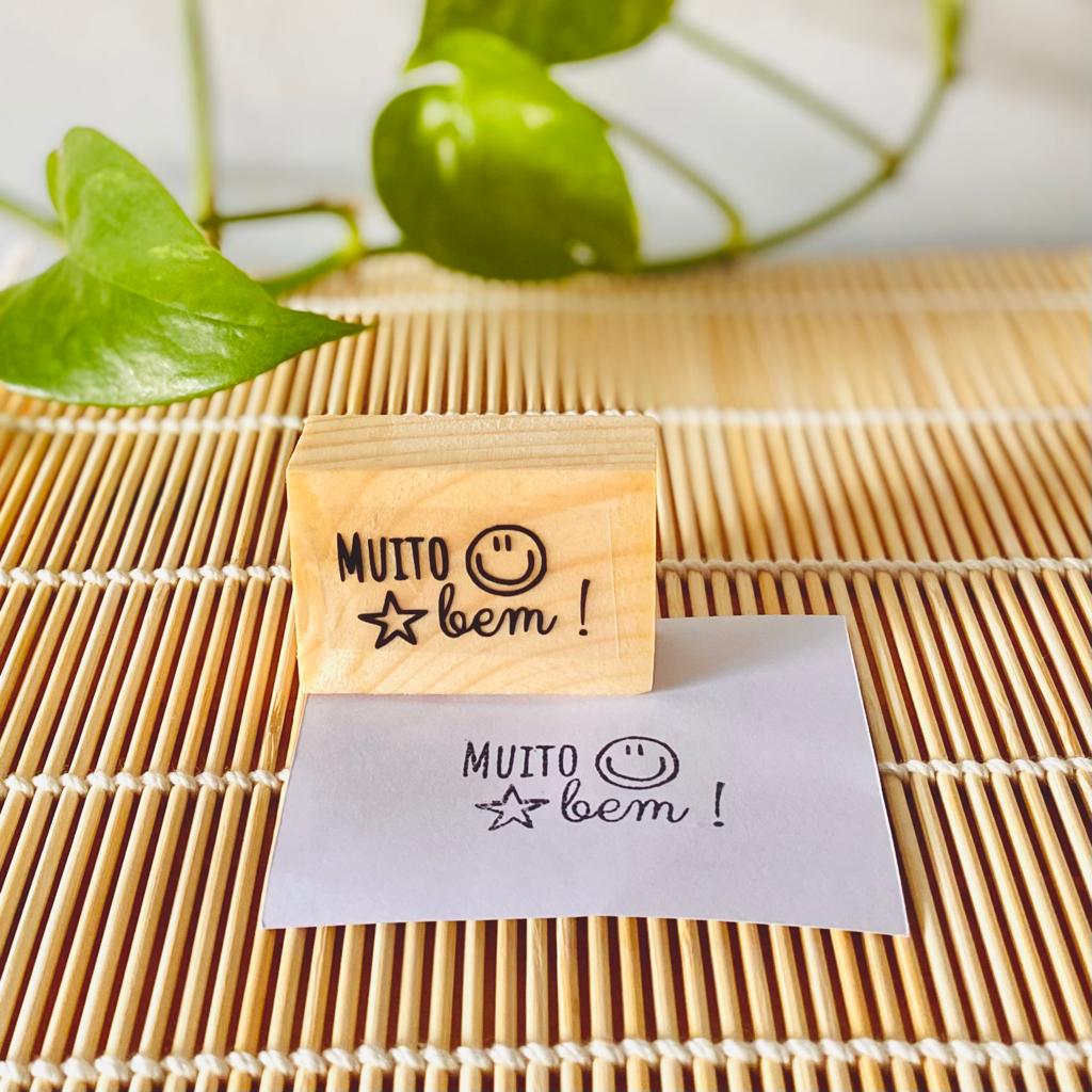 Carimbo de madeira com texto e desenho, e papel com impressão num tapete de vime.