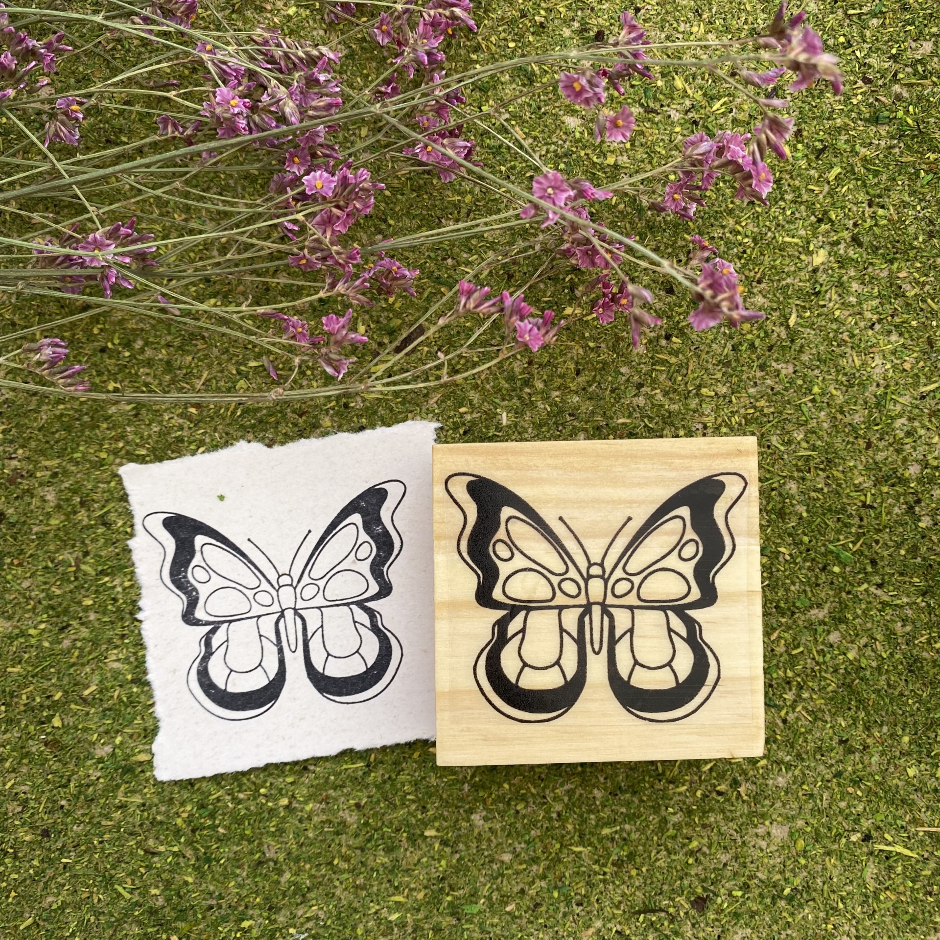 Carimbo de borboleta em madeira com padrão preto e impressão em papel sobre relva verde com flores lilases