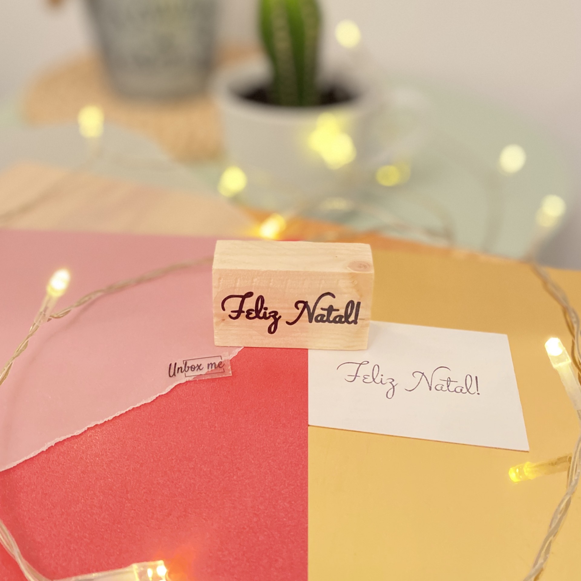Carimbo de madeira 'Feliz Natal!' sobre papéis coloridos e cartão branco com luzes decorativas