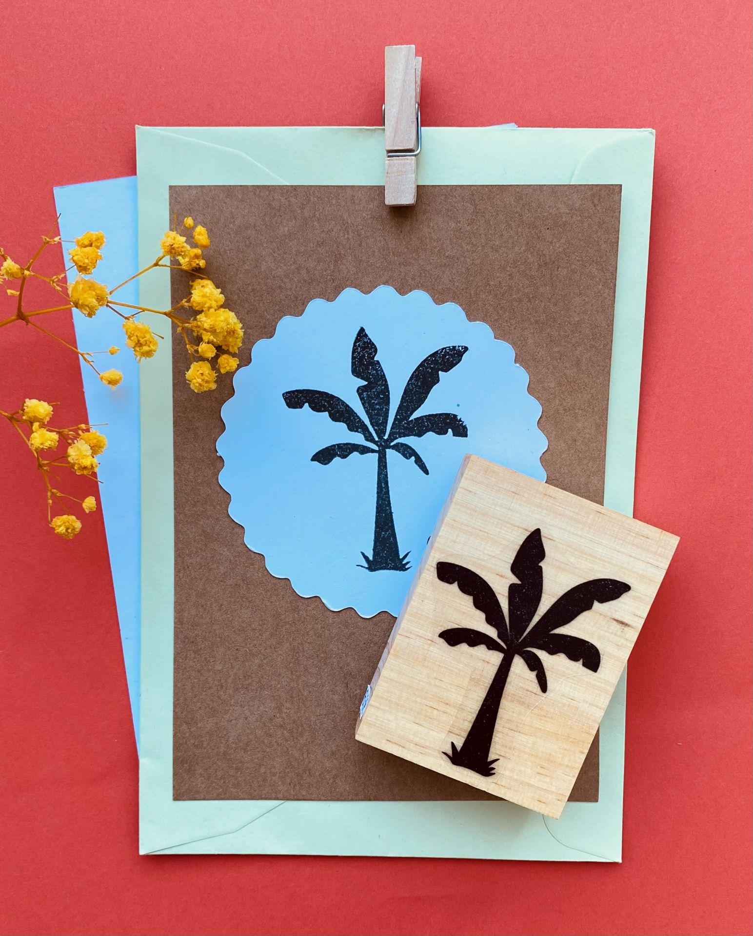 Carimbo de madeira com motivo de palmeira, cartão castanho com círculo azul, envelopes e flores amarelas num fundo vermelho.