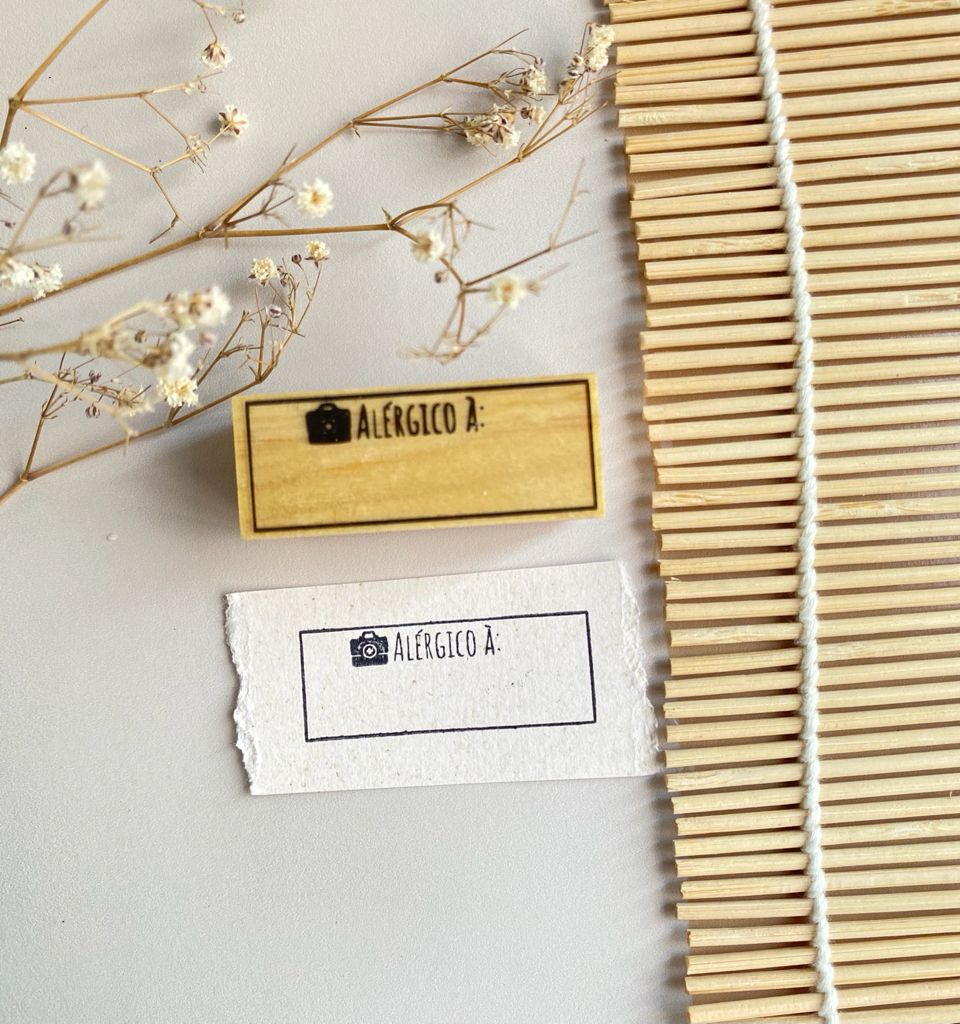 Dois carimbos 'ALÉRGICO À:' em madeira e papel, tapete de madeira e flores secas.