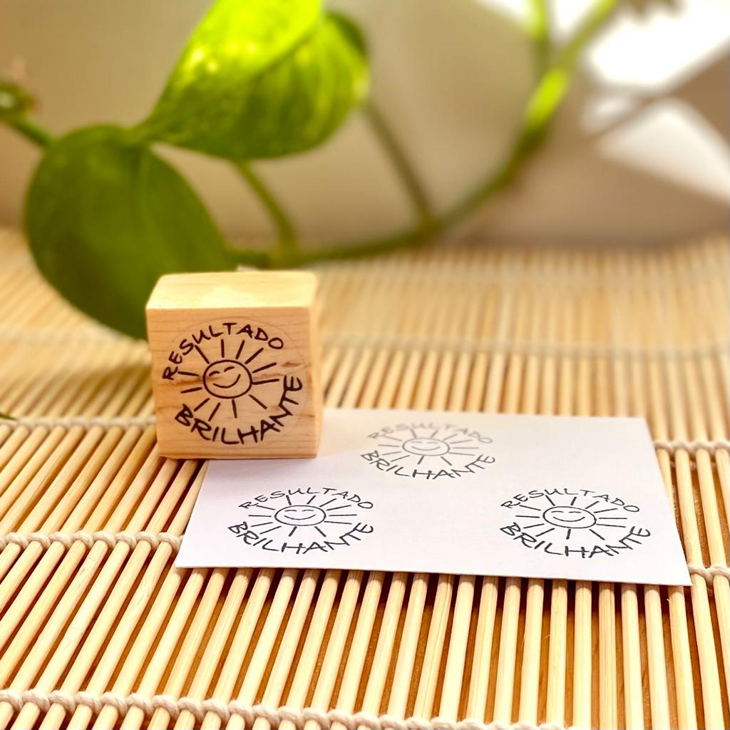 Carimbo de madeira com desenho de sol e texto 'RESULTADO BRILHANTE' sobre superfície de palha e papel estampado