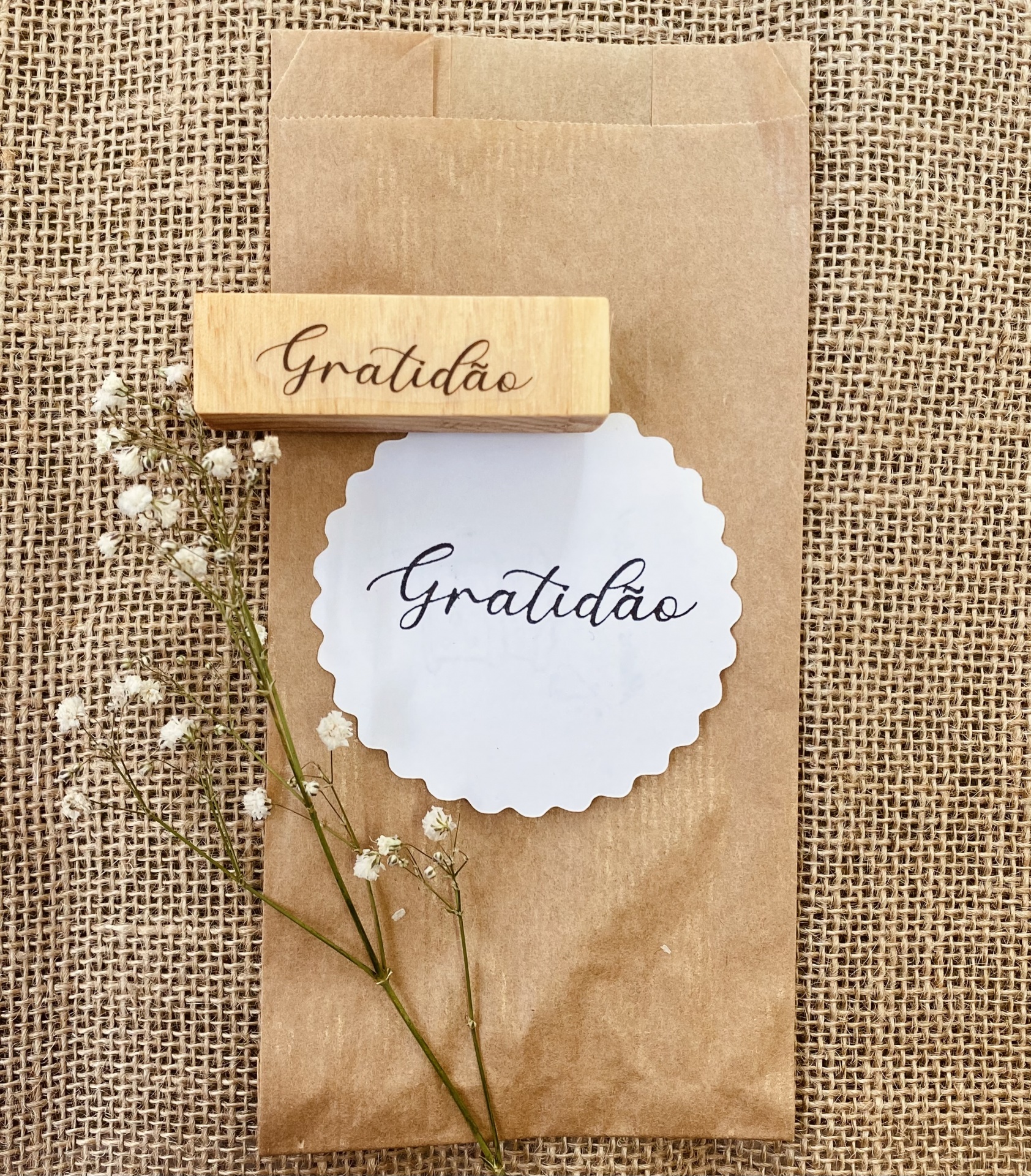 Carimbo de madeira, papel branco com 'Gratidão', saco de papel castanho e flores brancas sobre tecido arrojado.