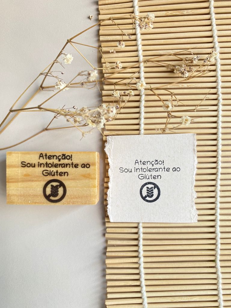 Carimbo de madeira e papel com texto Atenção! Sou intolerante ao Glúten ao lado de flores secas em tapete de bambu.