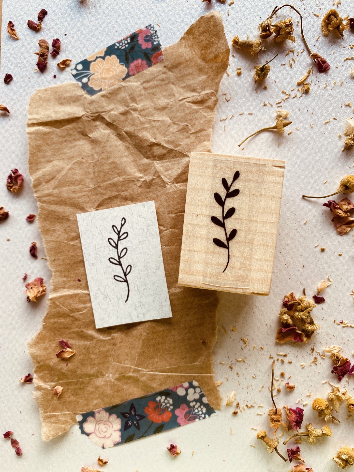 Carimbo de madeira com desenho de ramo de folhas e impressão em papel sobre papel castanho com fita decorativa e pétalas secas