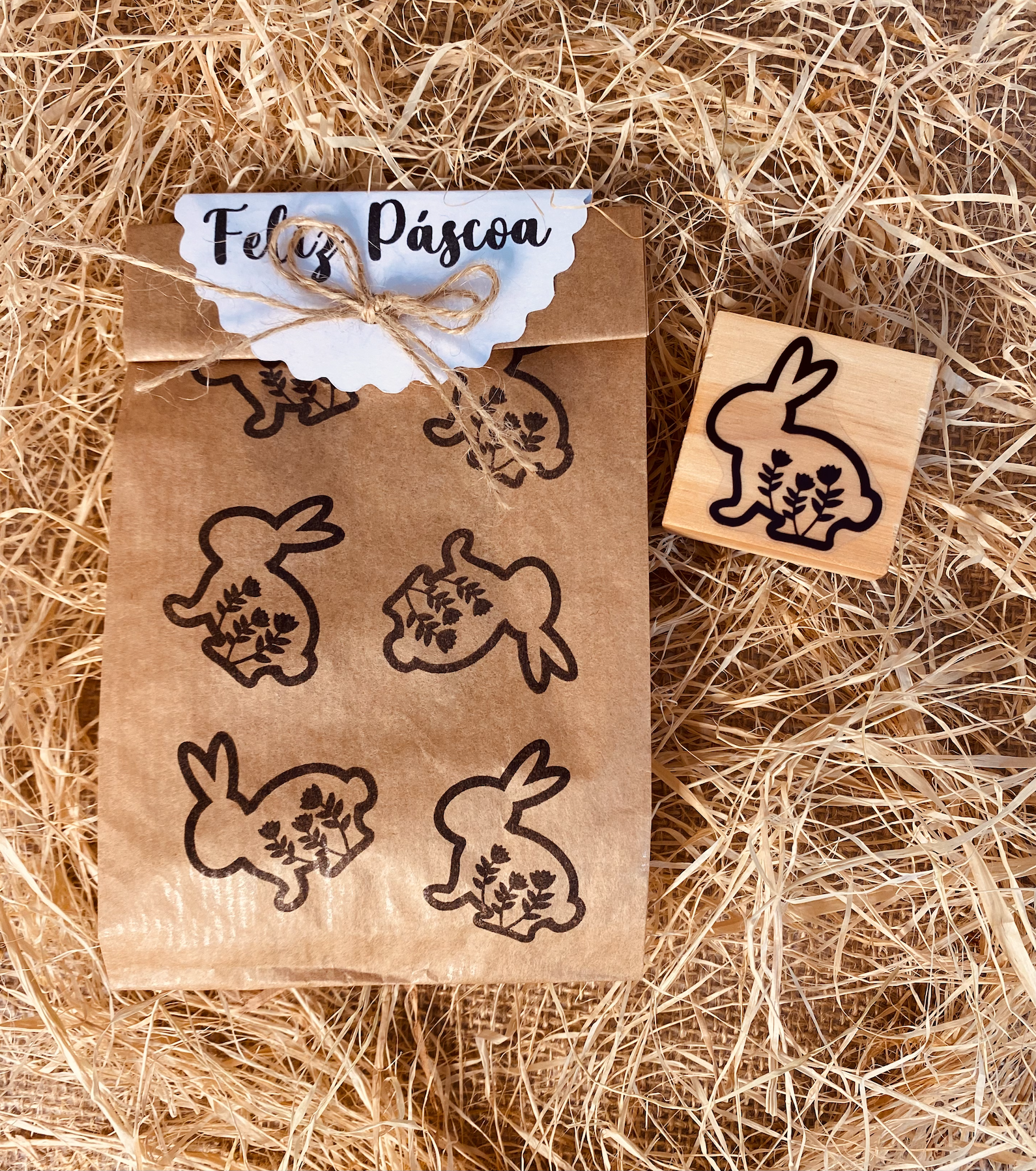 Saco de papel com estampados de coelhos e carimbo de madeira ao lado, sobre fundo de palha seca.
