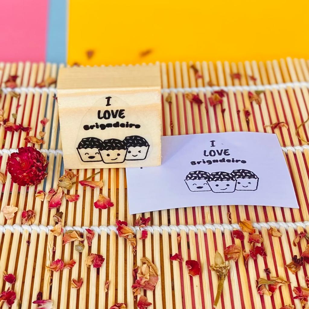 Carimbo de madeira com frase I LOVE brigadeiro e três rostos desenhados, sobre esteira de bambu com pétalas secas ao redor