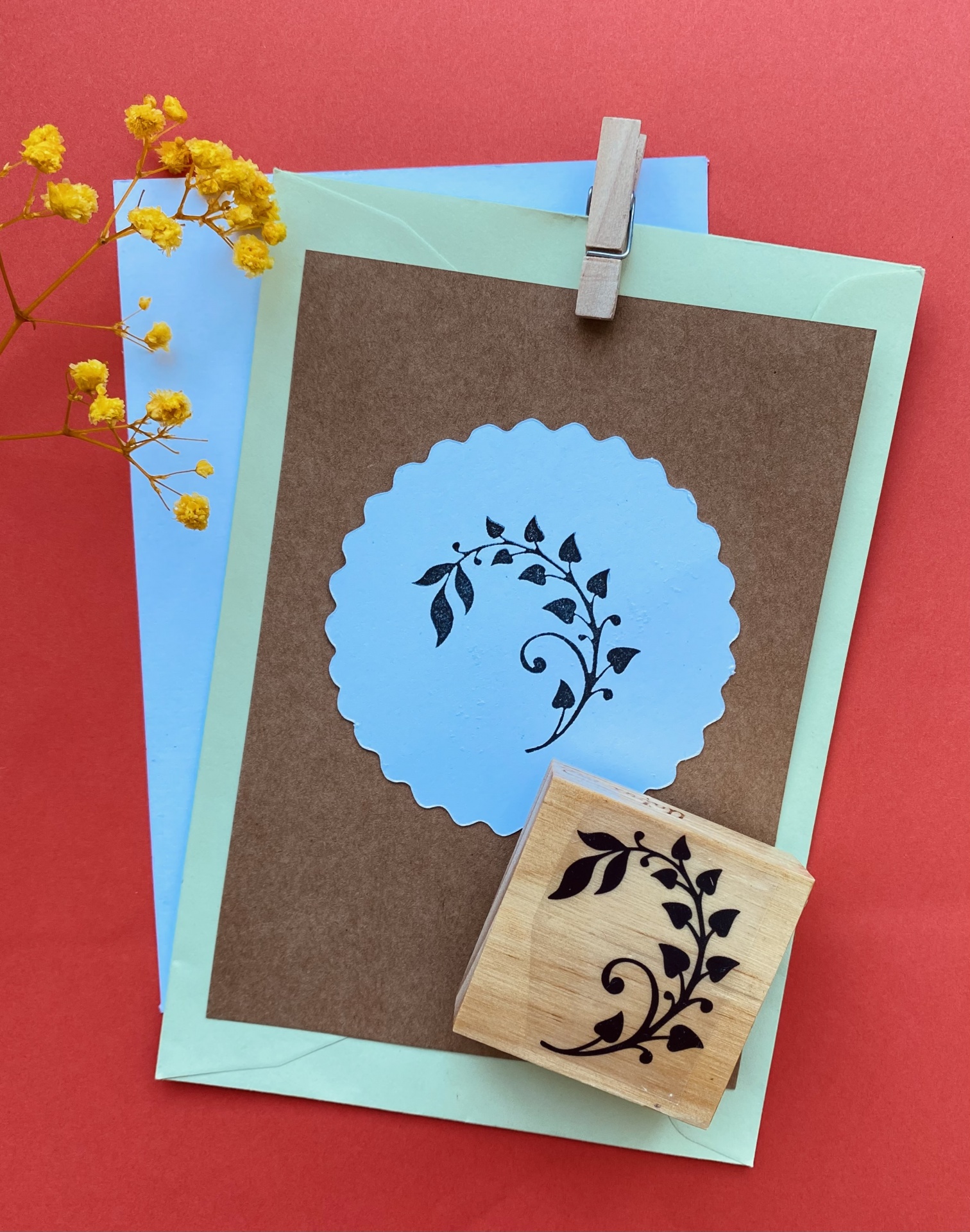 Carimbo de madeira com padrão floral, envelopes coloridos e cartão com recorte circular, flor amarela e fundo vermelho.