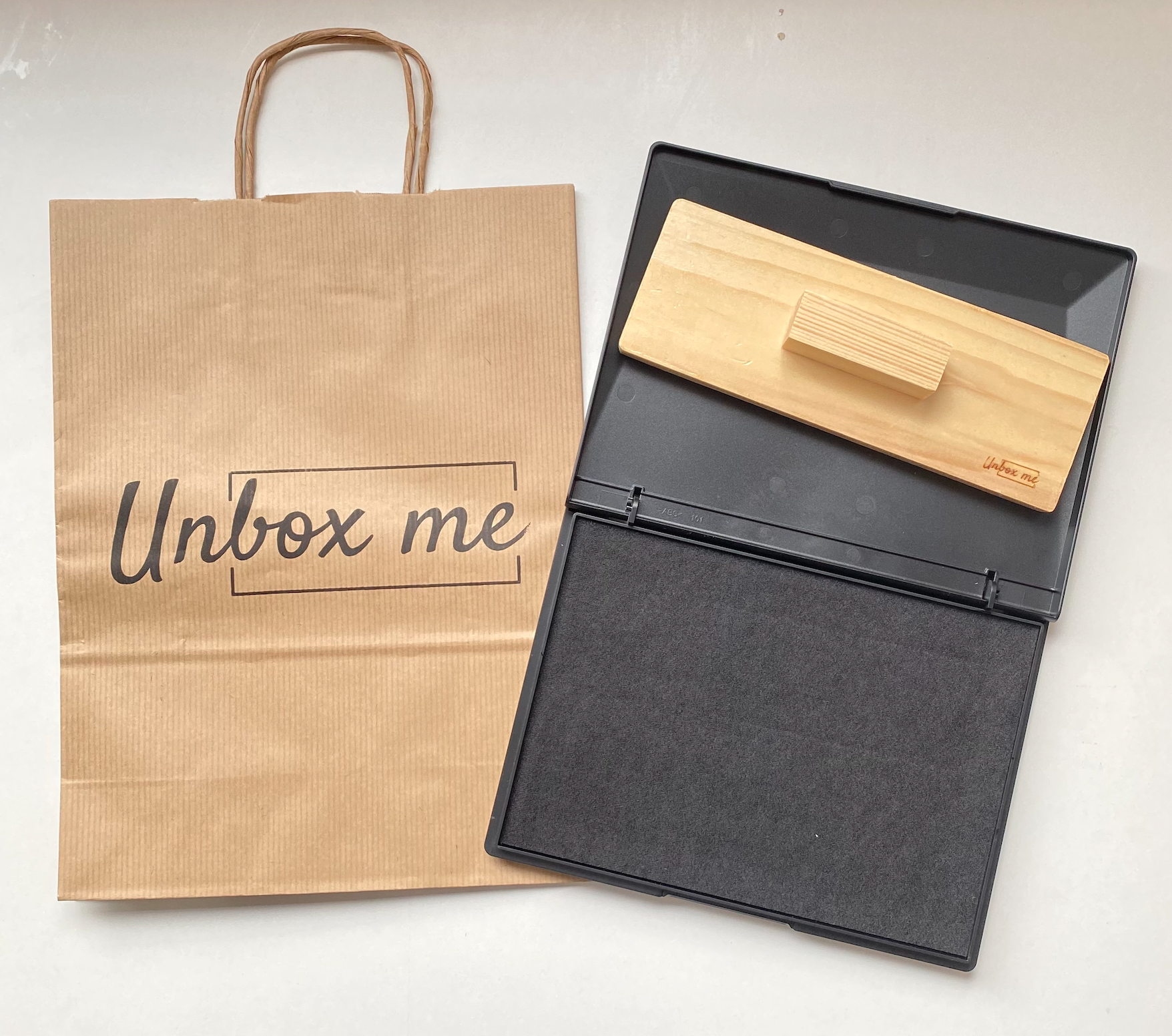Saco de papel com texto 'Unbox me' e caixa preta com base de madeira clara
