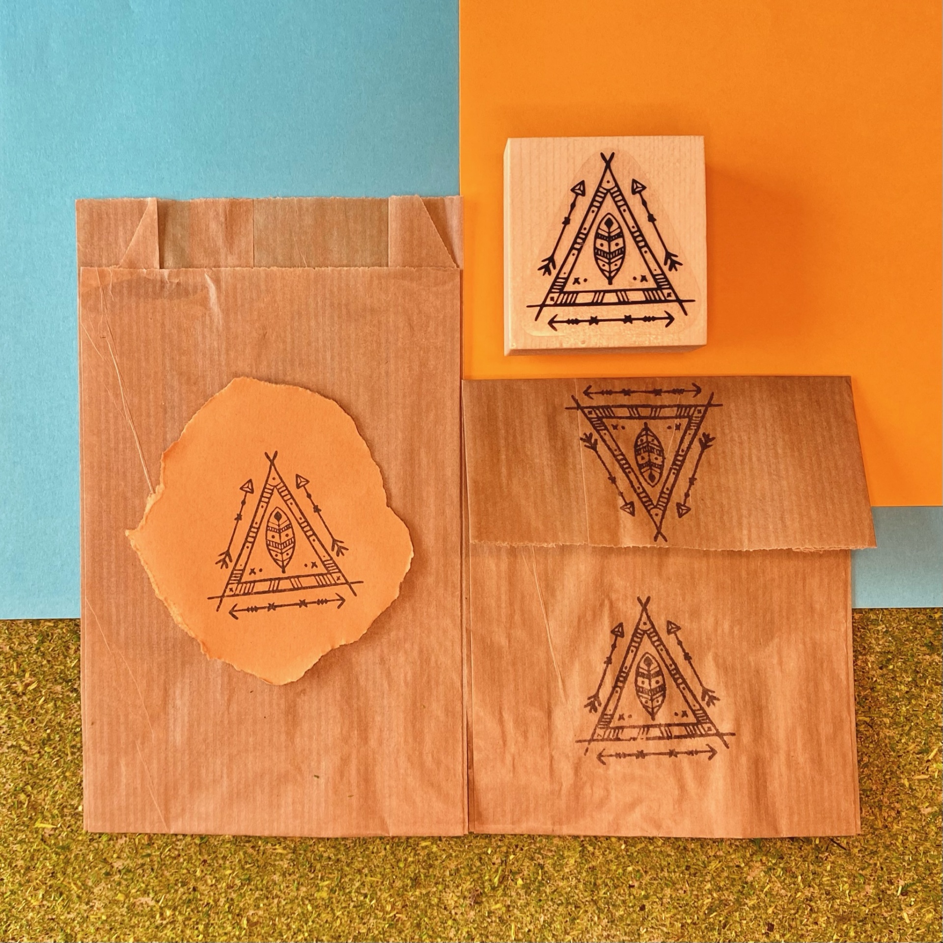 Sacolas de papel castanho com carimbo triangular preto em fundo azul e laranja.