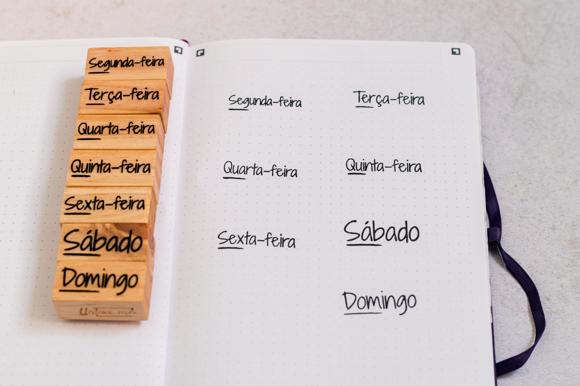 Caderno aberto com páginas pontilhadas e carimbos de madeira com nomes dos dias da semana em português.