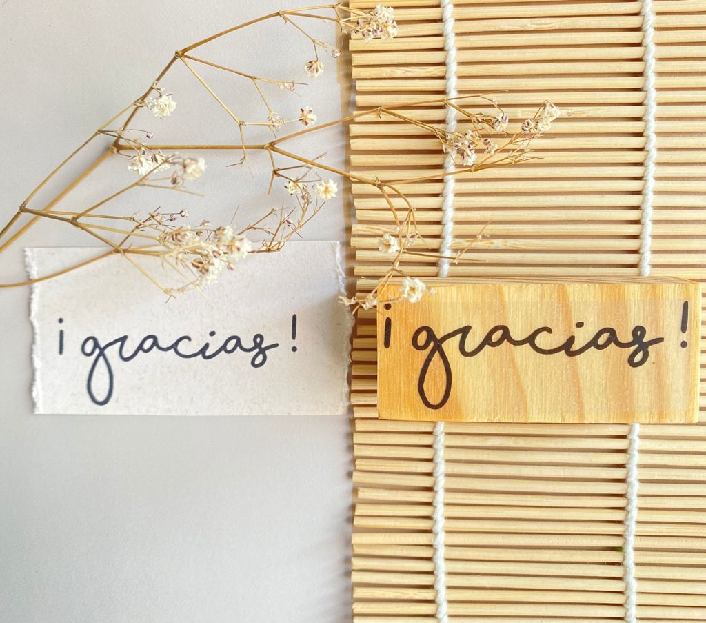 Placas de madeira clara e cartão branco com a palavra 'gracias!' escrita à mão, decoradas com ramos secos, sobre superfície clara e esteira de bambu.