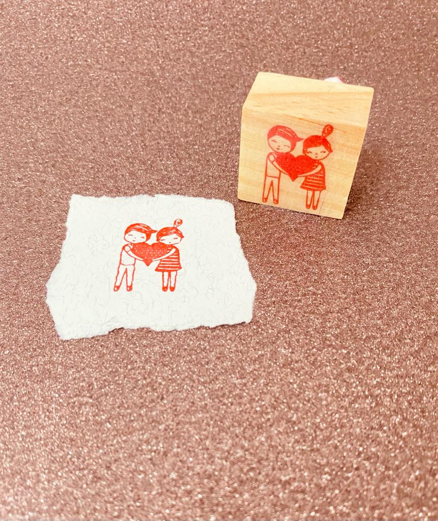 Carimbo de madeira com imagem de duas crianças segurando um coração vermelho e papel com carimbo.