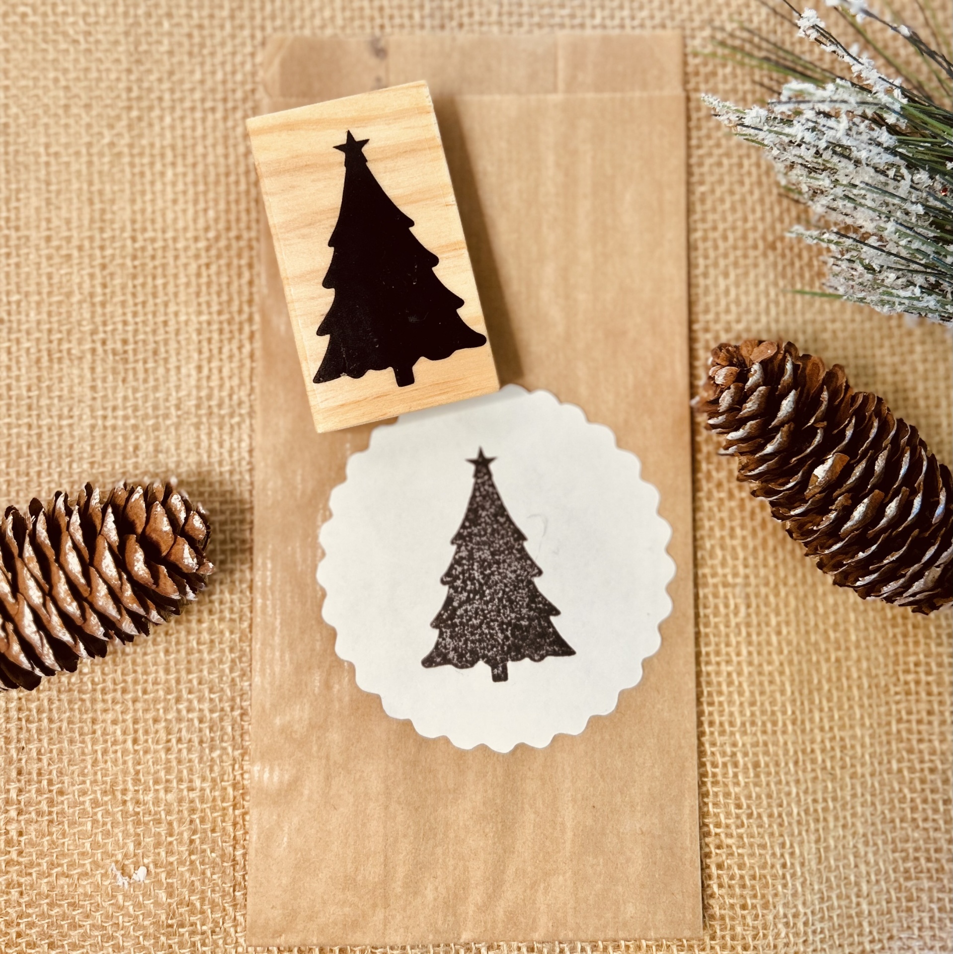 Carimbo de madeira com árvore de Natal preta e sachê de papel castanho com impressão do carimbo e pinhões