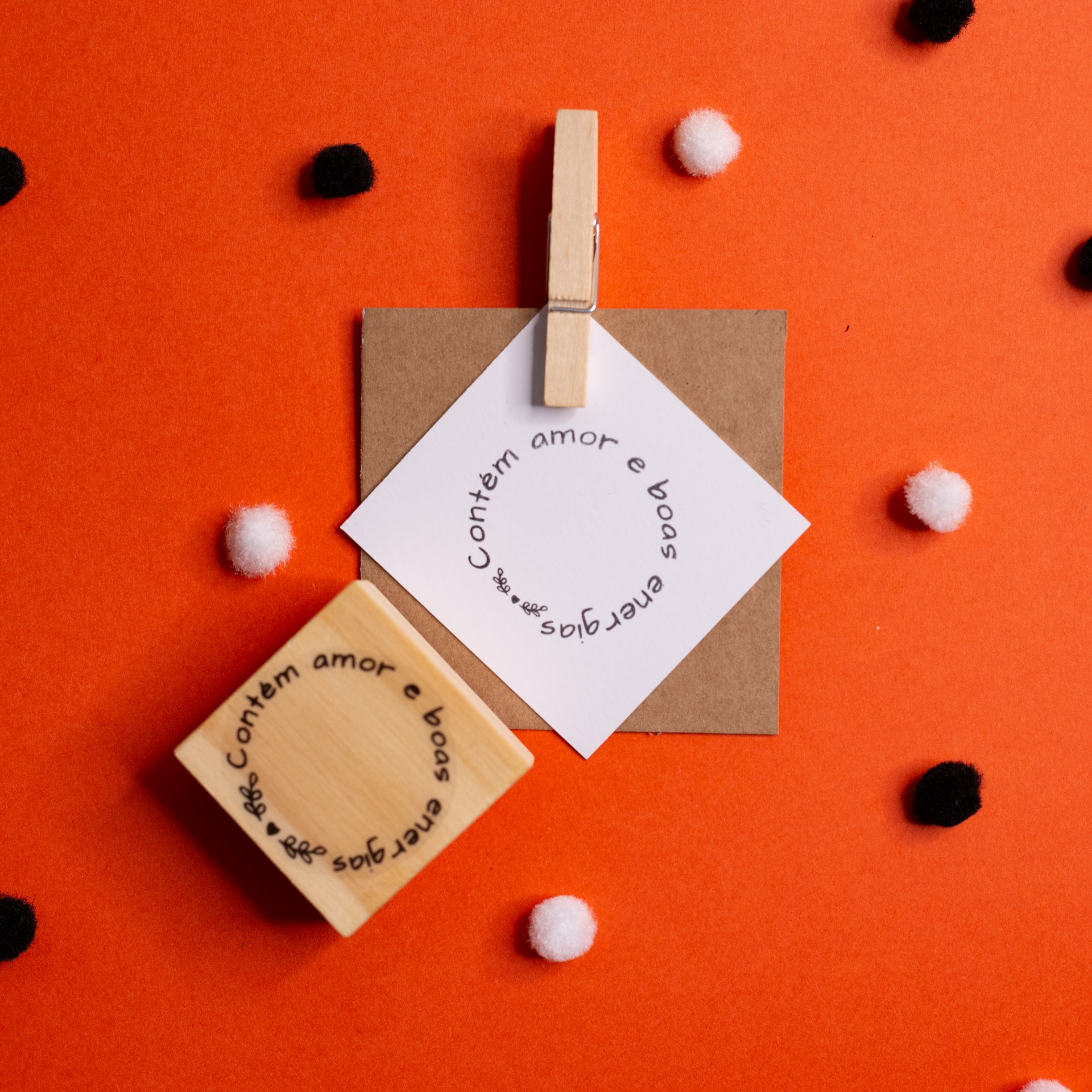 Carimbo de madeira com texto circular e selo branco similar com pregador sobre fundo laranja com pompons