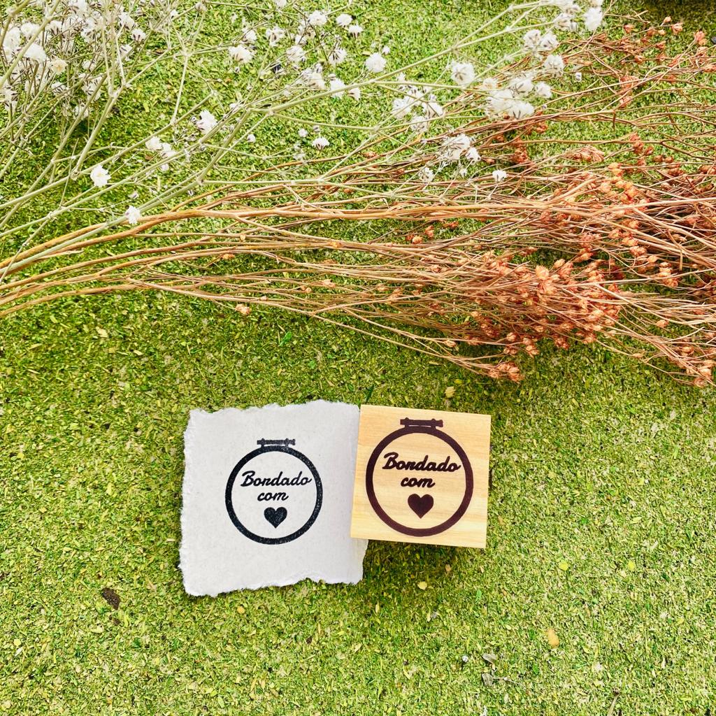 Carimbo quadrado de borracha com texto 'Bondado com' e coração sobre tecido branco e ramos secos em fundo verde