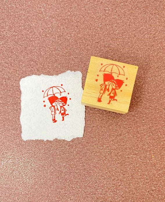 Carimbo de madeira com desenho de duas personagens e guarda-chuva, impressão em papel.