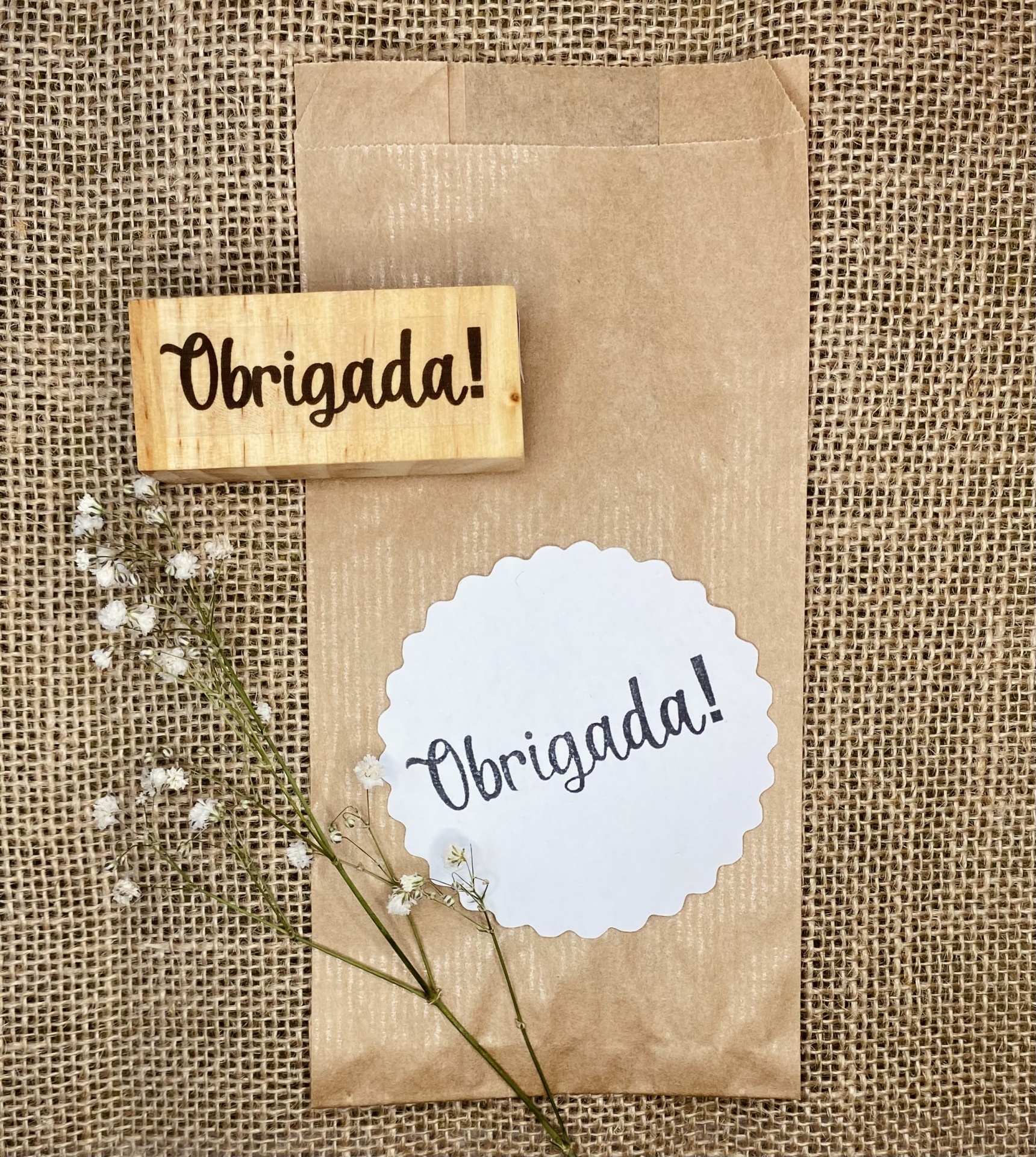 Saco de papel com etiqueta 'Obrigada!', carimbo de madeira e flores pequenas sobre fundo de juta