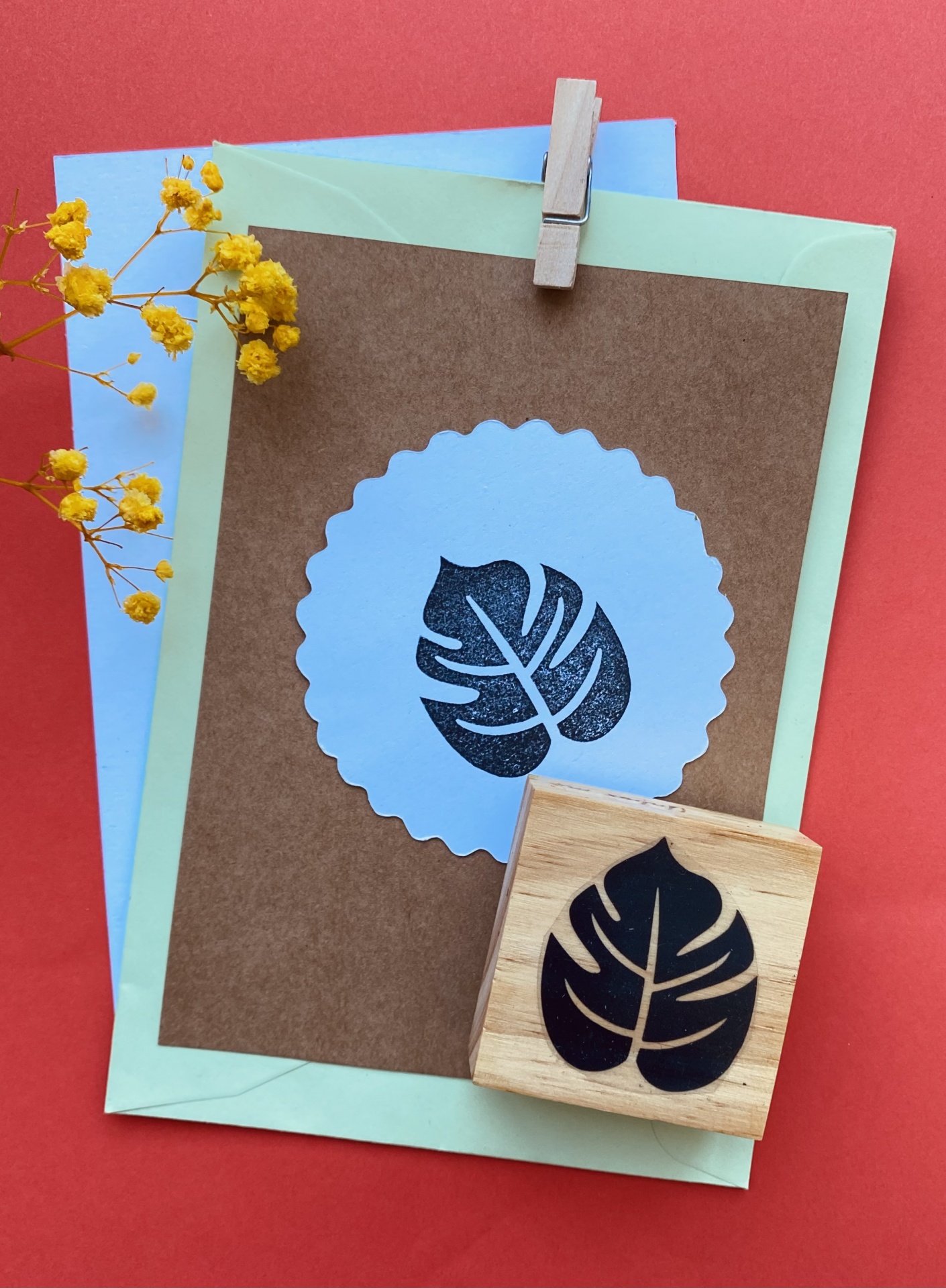 Cartão castanho com folha estampada preta em círculo branco, envelopes e flores amarelas secas sobre fundo vermelho.