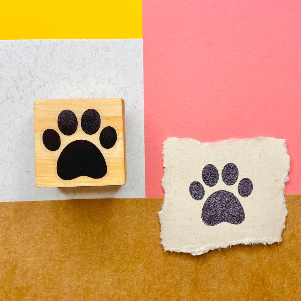 Carimbo de madeira com impressão preta de pata de animal ao lado da impressão numa peça de papel