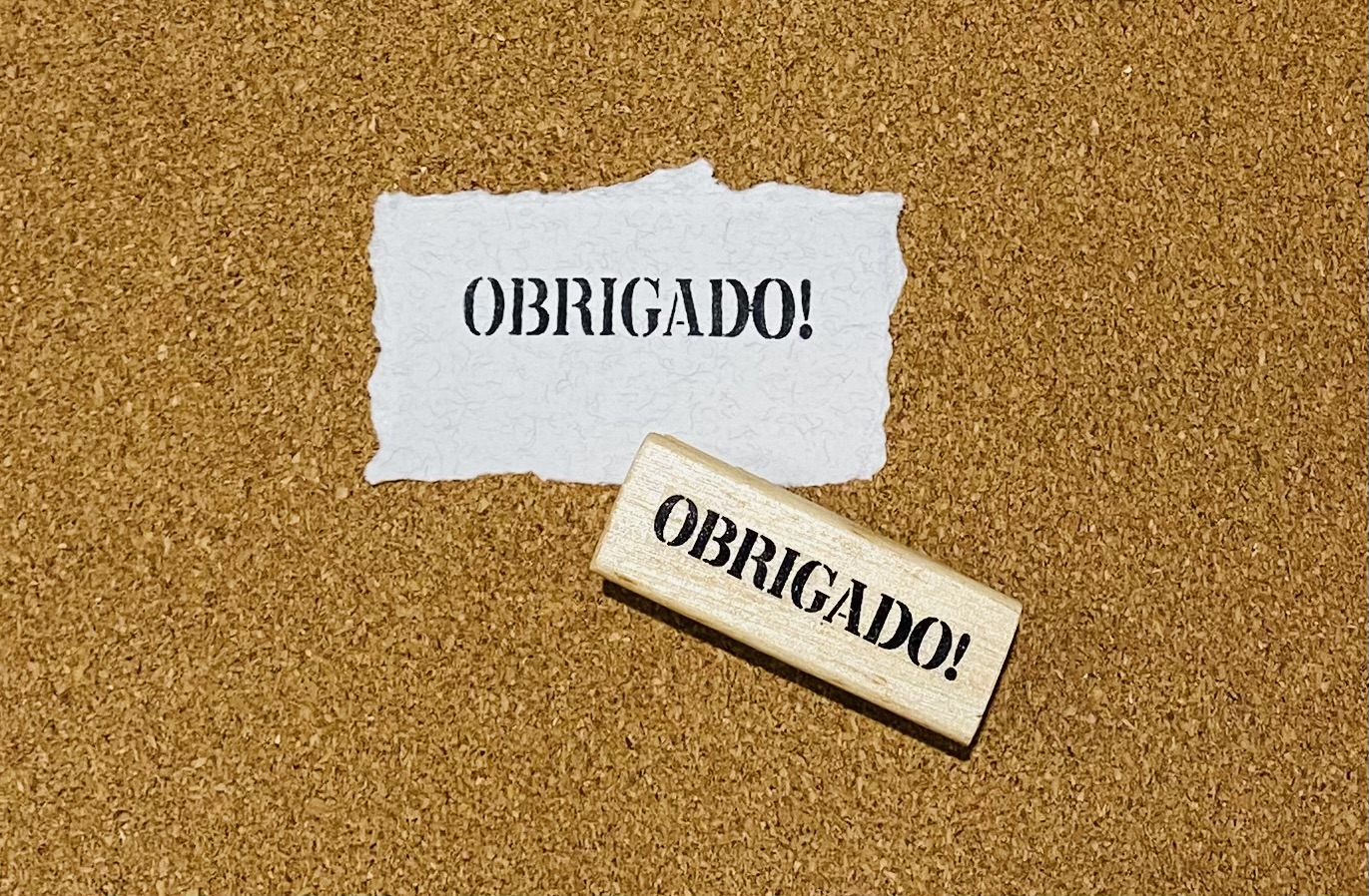 Carimbo de madeira com a palavra 'OBRIGADO!' e papel com a mesma palavra sobre cortiça