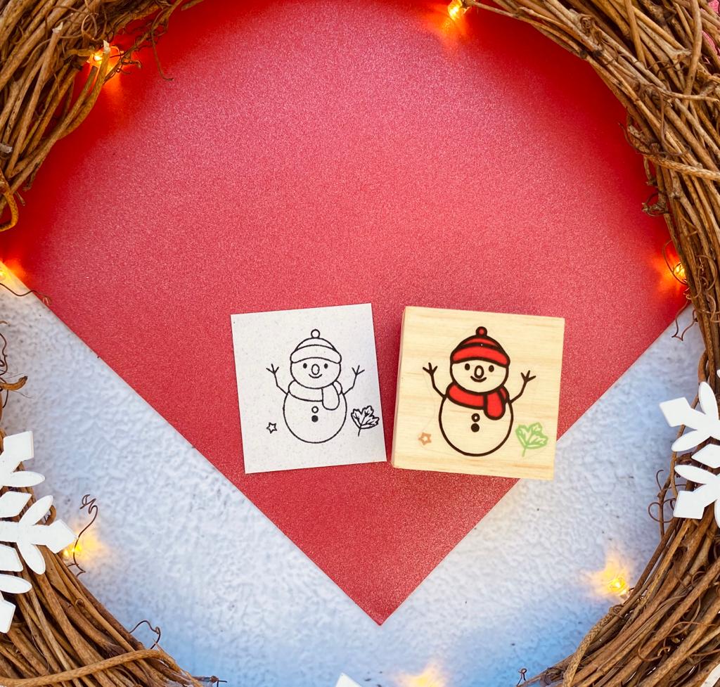 Dois carimbos de madeira com desenho de boneco de neve sobre fundo vermelho em forma de coração e guirlanda com luzes