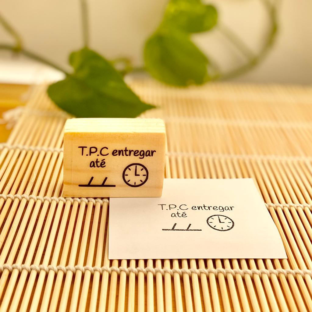 Carimbo de madeira com mensagem 'T.P.C entregar até' ao lado de folha carimbada sobre superfície de ripas de madeira