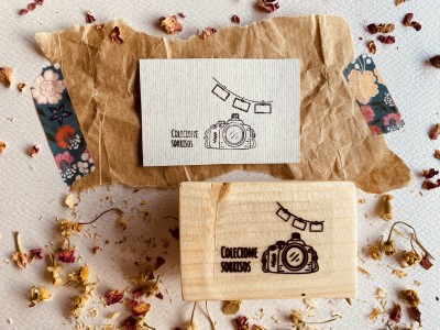 Carimbo de madeira com desenho de câmera e bandeirola, cartão com mesma imagem, papel castanho amassado e flores secas