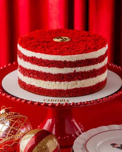 Bolo red velvet redondo com camadas alternadas de creme, em prato vermelho com ornamentos de Natal dourados e vermelhos ao redor.
