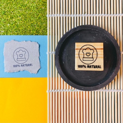 Bloco de madeira com carimbo 100% NATURAL em esteira de bambu ao lado de papel com mesma imagem e fundo colorido.