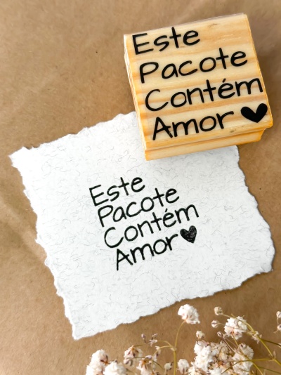 Carimbo de madeira com frase 'Este Pacote Contém Amor' e papel branco com mesma frase sobre fundo castanho e flores secas