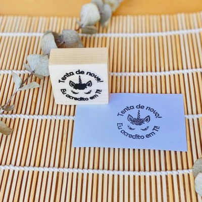 Carimbo em madeira com texto motivacional e desenho de unicórnio colocado sobre papel azul e esteira de madeira