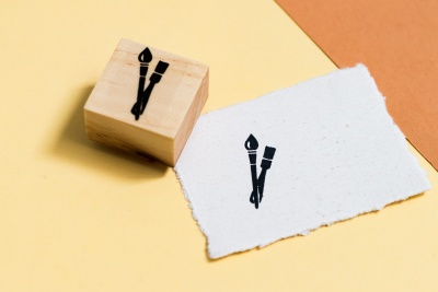 Carimbo de madeira quadrado com desenho de pincéis cruzados e papel branco com o mesmo desenho sobre fundo amarelo