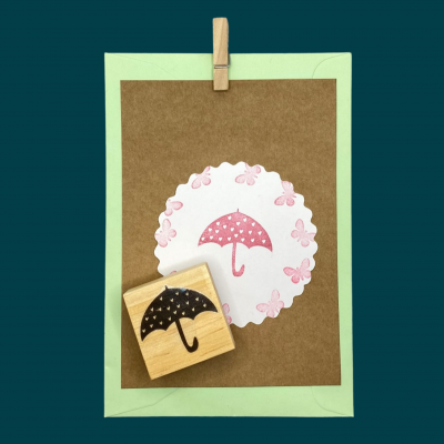 Carimbo de guarda-chuva preto sobre papel decorativo com guarda-chuva rosa e flores cor-de-rosa, em suporte verde claro com pregador de madeira