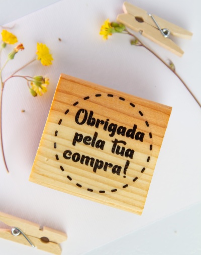 Bloco de madeira com mensagem gravada 'Obrigada pela tua compra!' sobre fundo branco com flores amarelas e prendedores de madeira