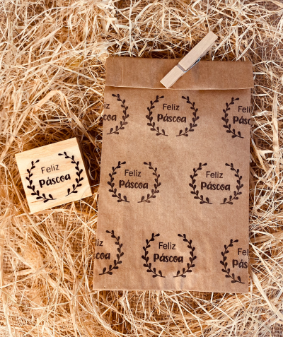 Selo de borracha com carimbo e saco de papel com impressão 'Feliz Páscoa' em tinta preta sobre fundo de palha.