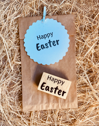 Saco de papel com etiqueta azul e carimbo 'Happy Easter' sobre palha