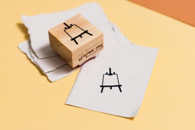 Carimbo de madeira com desenho de cavalete e duas folhas de papel impressas.