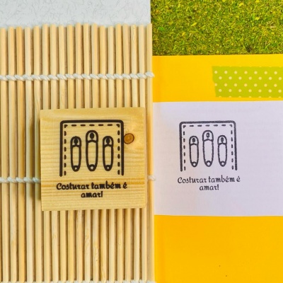 Carimbo de madeira com ícones de alfinetes e texto Costurar também é amar! ao lado de impressão do carimbo em papel amarelo