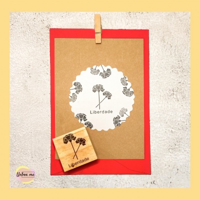 Cartão kraft com envelope vermelho, etiqueta branca com flores e texto 'Liberdade' e carimbo de madeira
