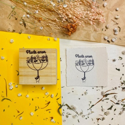 Selo de madeira com estampa 'Plante amor' ao lado de papel branco com a mesma estampa, em fundos amarelo e branco com flores secas.