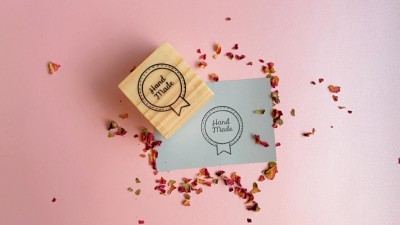 Selo de madeira com texto 'Hand Made' sobre fundo rosa com papel cinzento e pétalas secas