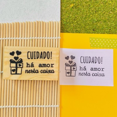 Dois cartões com a mensagem 'CUIDADO! há amor nesta caixa' e desenho de uma caixa com corações, um em madeira e outro em papel, sobre fundos amarelo e varetas de madeira.
