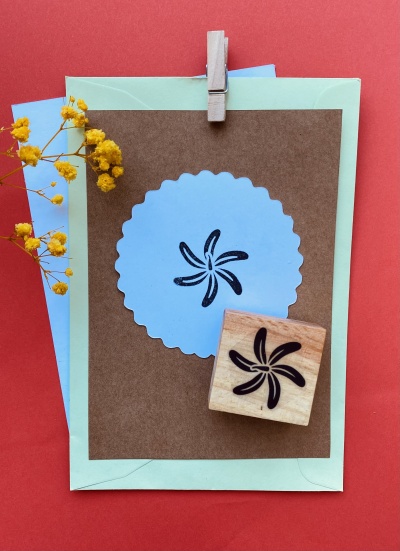 Carimbo de madeira com desenho em espiral preto sobre cartão castanho e etiqueta azul, envelopes e flores amarelas