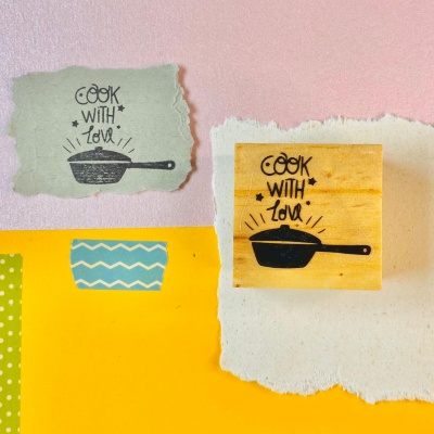 Selo de borracha amarelo com desenho de frigideira e texto 'COOK WITH LOVE' sobre fundo colorido