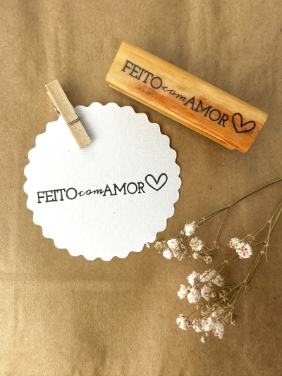 Carimbo de madeira com texto 'FEITO com AMOR' e etiqueta branca com o mesmo texto, com flores secas ao lado.