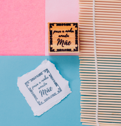 Peça de madeira com carimbo e papel com frase para mãe sobre superfícies coloridas e esteira de bambu
