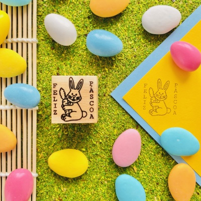 Carimbo de madeira com coelho e texto FELIZ PASCOA ao lado de ovos coloridos sobre grama.